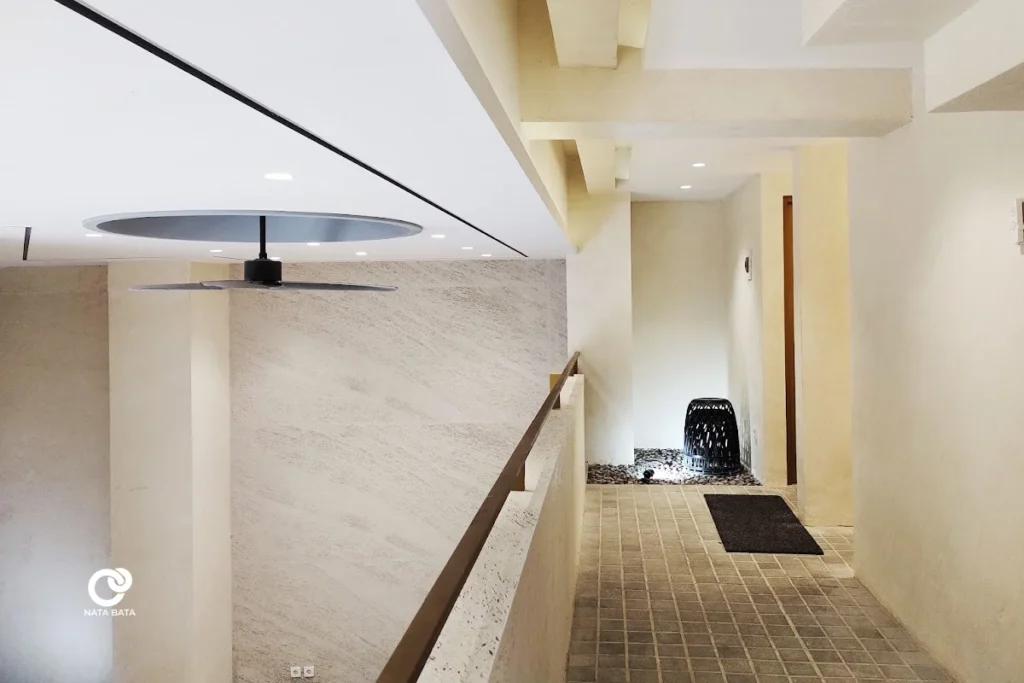 Lorong menuju kamar mandi lantai dua dengan pencahayaan natural dan elemen batu koral, desain bersih dan hangat.